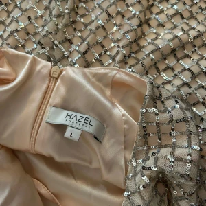 Champagnefärgad långklänning Hazel - Snygg långklänning från Hazel i champagnefärgad satin med glittriga paljetter i rutmönster. Klänningen har smala axelband med snörning i ryggen och en slits framtill. Perfekt för dig som vill sticka ut med en elegant och glittrig look.