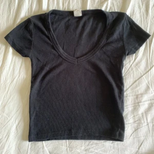 Svart ribbad croppad topp - En svart, ribbad croppad topp med djup v-ringning och korta ärmar. Enkel och stilren design som passar perfekt till jeans eller kjol. Mjuk och stretchig bomullskänsla för en bekväm passform.