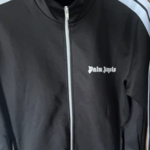 Svart Palm Angels - Svart vindjacka från Palm Angels med vit dragkedja och logga på bröstet. Jackan har hög krage och raka linjer, perfekt för en clean och sportig look. Materialet känns lätt och smidigt, och den har en klassisk passform.
