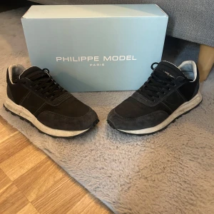 Svarta sneakers från Philippe Model - Snygga svarta sneakers från Philippe Model med vita sulor och svarta snören. Skorna har en klassisk design med rund tå och är tillverkade i mocka och syntet. Perfekta för dig som gillar stilrena och bekväma skor. TVÄTTAS INANN!!!
