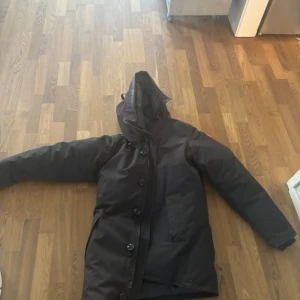 Äkta Canada Goose - Säljer en svart dunjacka från Canada Goose med huva och knäppning framtill. På jackans luva finns det tecken av andvändning.Jackan har en klassisk look med stora fickor och diskret logga på ärmen. Perfekt för kalla dagar när du vill hålla dig varm och stilsäker. Självklart äkta.Det är bara att skicka bud. Och såklart, fråga på! 