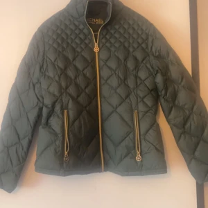 Grön quiltad jacka från Michael Kors - Snygg grön quiltad dunjacka från Michael Kors med guldfärgade dragkedjor och MK-detaljer. Jackan har lång ärm och hög krage, perfekt för kyligare dagar. Klassisk och stilren design som passar till många tillfällen. 