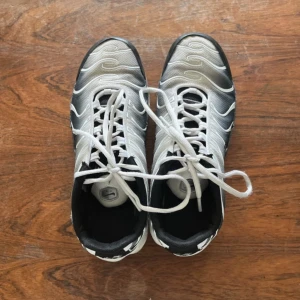Nike Air Max Plus Tuned 1 sneakers svart/vit - Nike Air Max Plus Tuned 1 sneakers i svart och vitt med vita snören och synliga luftbubblor i sulan. Skorna har vågiga detaljer på ovandelen och en robust, sportig design. Perfekta för dig som gillar streetwear och klassiska Nike-modeller.