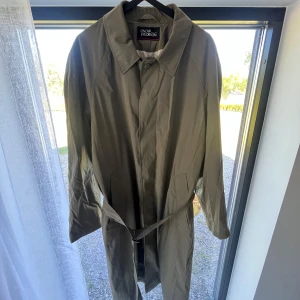 Khaki trenchcoat från Oscar Jacobson - Stilren olivgrön trenchcoat från Oscar Jacobson med klassisk krage och knäppning framtill. Jackan har bälte i midjan och innerficka med dragkedja. Tillverkad i bomull och polyamid för en lätt och bekväm känsla. C96 i storlek men passarM-L beroende på hur man vill ha passformen. Mått på begäran