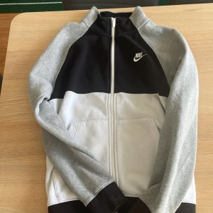 Nike ziptröja  - Säljer en snygg ziptröja från Nike i färgerna svart, grå och vit. Tröjan har dragkedja framtill, två fickor och klassisk Nike-logga på bröstet. Perfekt för en sportig look eller till vardags.