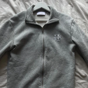 Grå zipup -kofta från Les Deux - Snygg grå zip-jacka från Les Deux med broderad logga på bröstet. Koftan har hög krage och dragkedja framtill. Perfekt för lager-på-lager och enkel att matcha med olika outfits.😮‍💨Säljer så min bror inte använder den längre, precis som ny🤩‼️