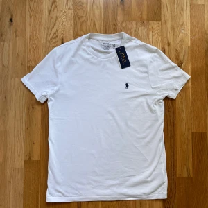 Vit t-shirt från Polo Ralph Lauren - HELT OANVÄND. Klassisk vit t-shirt från Polo Ralph Lauren med blå broderad logga på bröstet. Modellen är slim fit och har rund hals och korta ärmar.