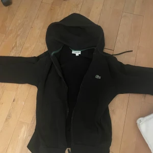 Svart hoodie från Lacoste - Säljer en svart hoodie från Lacoste med dragkedja och huva. Klassisk krokodillogga på bröstet och två fickor framtill. Perfekt för en avslappnad stil och enkel att matcha med det mesta.