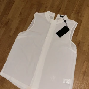 Vit ärmlös blus från Stockh LM - Säljer en stilren vit ärmlös blus från Stockh LM. Blusen har hög krage och är tillverkad i ett tunt, lätt material som ger en luftig känsla. Perfekt att matcha med både jeans och kjol.Den har en liten fläck trots att den är oanvänd men går inte att märka om man ej kollar noggrant.