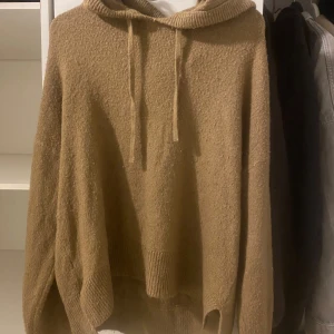 Beige stickad hoodie från Vero Moda - Mysig beige stickad hoodie från Vero Moda med huva och dragsko. Tröjan har långa ärmar och ribbade muddar vid ärmslut och nederkant. Perfekt för kyliga dagar och enkel att matcha med jeans eller leggings.