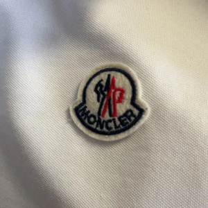 Vit piké t-shirt från Moncler - Säljer en klassisk vit piké t-shirt från Moncler med broderad logga på bröstet. T-shirten har krage, knappar framtill och korta ärmar med diskreta ränder på ärmsluten. Perfekt för en stilren look.