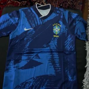 Blå Brasilien fotbollströja Nike - Säljer en blå Brasilien fotbollströja från Nike med abstrakt mönster och gula detaljer. Tröjan har korta ärmar, rund hals och Brasilien-emblem på bröstet. Perfekt för dig som vill visa support för landslaget!