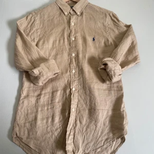 Beige linneskjorta från Ralph Lauren - Säljer en beige långärmad skjorta i linne från Ralph Lauren. Slim fit-modell med klassisk krage, knappar framtill och blå logga på bröstet. Perfekt till sommaren för en stilren och avslappnad look.