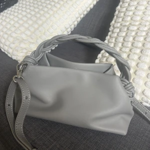 Grå axelväska med flätat handtag - Säljer en stilren grå axelväska med ett snyggt flätat handtag och justerbar axelrem. Väskan är gjord i syntetmaterial och har ett rymligt huvudfack. Perfekt för dig som vill ha en trendig accessoar till din outfit. Banden går att ta av ifall man skulle önska det. Använd 1-2 gånger så i nyskick. Pris kan diskuteras 