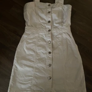 Vit jeansklänning med knappar från NA-KD - Säljer en vit jeansklänning från NA-KD i storlek XS. Klänningen har breda axelband, knappar framtill och två fickor vid höfterna. Perfekt för en trendig och avslappnad look. Jag har burit den en gång så den är i princip ny🌸🤍💕