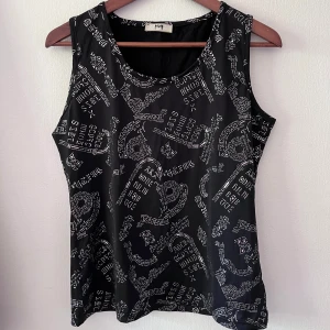Linne 90-tal - Svart ärmlös topp/bredaxlat linne från Joy med grafiskt mönster i silver. Strl L, stretchig polyesterblandning med rund halsringning. Snygg att ha över långärmad tröja och styla med jeans eller kjol. Superbra skick!