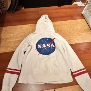 Vit NASA hoodie från H&M - Vit hoodie från H&M med stor NASA-logga framtill och röda ränder på ärmarna. Tröjan har huva och magficka, perfekt för dig som gillar rymden och vill ha en cool streetstil.