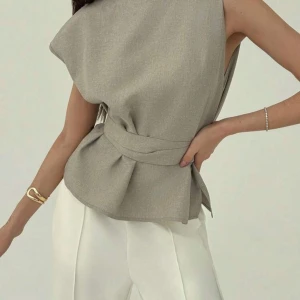 Beige omlottopp med knytband - Säljer en stilren beige omlottopp med knytband i midjan och lös passform. Toppen har ärmlös design och rund halsringning, perfekt för en chic och modern look.