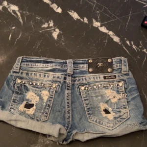 Miss Me jeansshorts med stjärnor och slitningar - Säljer ett par blå jeansshorts från Miss Me med coola slitningar, nitar och stjärnmönster på framsidan. Shortsen har uppvikta ben och detaljerade bakfickor med nitar och broderi. Perfekta för sommaren!