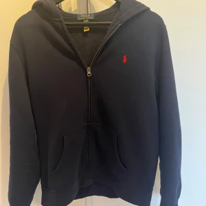 Mörkblå hoodie från Polo Ralph Lauren - Säljer en mörkblå hoodie från Polo Ralph Lauren med dragkedja och huva knappt använd Tröjan har en röd broderad logga på bröstet och två fickor framtill. Perfekt för en avslappnad stil. Barn Stl 18-20 men sitter som xs/s