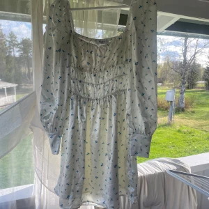 Blommig långärmad klänning från NA-KD - Supersöt vit klänning med blått blommönster från NA-KD. Klänningen har långa puffärmar med knappar vid ärmslut, smockad rygg och markerad midja. Perfekt för vår och sommar, med romantisk och feminin känsla.