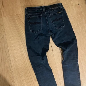 Nudie Jeans 29/32 - Säljer ett par mörkblå jeans från Nudie Jeans med klassisk femficksdesign och smal passform. Byxorna har snygga detaljer på bakfickorna och silverfärgade knappar.