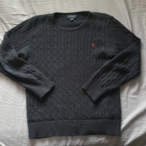 Mörkblå kabelstickad tröja från Polo Ralph Lauren - Kabelstickad svart tröja från Polo Ralph Lauren med klassisk röd broderad logga på bröstet. Tröjan har rund halsringning och långa ärmar med ribbade muddar och nederkant. Perfekt för en stilren och tidlös look.