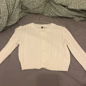 Vit ribbad kofta från H&M - Säljer en söt vit kofta från H&M med ribbat mönster och rund halsringning. Koftan är croppad och har långa ärmar samt knappar framtill. Perfekt att slänga över en topp för en enkel och stilren look.