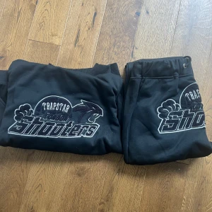 Svart set från Trapstar Shooters - Svart set från Trapstar Shooters med broderad logga och text på bröstet. Mjuk insida och ribbade muddar vid ärmslut och nederkant. Perfekt för en avslappnad streetwear-look. Säljer pga fel storlek 