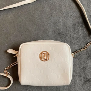 Vit axelväska från River Island - Säljer en stilren vit axelväska från River Island med guldfärgad kedja och logga framtill. Väskan har präglat mönster med RI-loggor och tillhörande axelrem. 🤍
