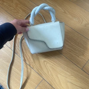 Vit handväska från ATP Atelier - Säljer en stilren vit handväska från ATP Atelier med unik asymmetrisk lockdesign och tvinnat handtag. Väskan har axelrem och är tillverkad i skinn. Perfekt accessoar för att lyfta din outfit! Ganska sliten, se bild!