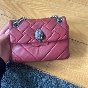 Röd Kurt geiger axelväska med kedja - Säljer en röd Kurt geiger axelväska med silverfärgad kedja och dekorativt spänne framtill. Väskan har ett huvudfack och är tillverkad i skinnliknande material. Perfekt för dig som vill ha en snygg accessoar med cool detalj. Den är ganska sliten, se bild!