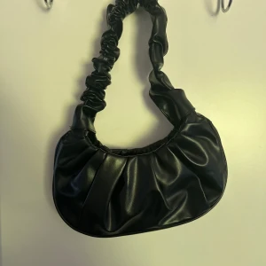 Svart axelväska med rynkad rem - Säljer en svart axelväska i syntetmaterial med snygg rynkad rem och rundad form. Perfekt storlek för det viktigaste och passar till många olika outfits. Enkel men trendig design.