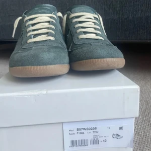 Gröna sneakers från Maison Margiela - Säljer ett par gröna sneakers från Maison Margiela med beige snörning och gummisula. Skorna har en mix av mocka och skinn, klassisk design och låg profil. Perfekta för dig som gillar stilrena och unika sneakers.