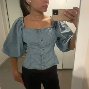 Ljusblå jeansblus med puffärmar - Trendig ljusblå blus i jeanstyg med stora puffärmar och knappar framtill. Fyrkantig ringning och figurnära passform ger en snygg siluett. Skriv för frågor!