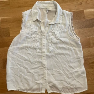 Vit ärmlös blus med spetsdetaljer från Divided - Säljer en vit ärmlös blus från Divided med fina detaljer och spetsbroderier framtill. Blusen har krage och perldetaljer till knäppning framtill, perfekt för en somrig look.