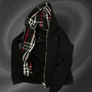 Burberry Hoodie  - Säljer en svart hoodie med dragkedja från Burberry. Insidan av huvan har ett klassiskt rutigt mönster i svart, vitt och rött. Tröjan har två fickor framtill och justerbar huva med snören.