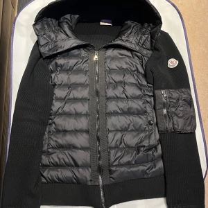 Svart Cardigan från Moncler - Säljer en svart Moncler-jacka med quiltad front och huva. Jackan har stickade ärmar och sidor, dragkedja framtill och en ficka med dragkedja på ärmen. Klassisk Moncler-logga på ärmen. Perfekt för lager-på-lager under kallare dagar.