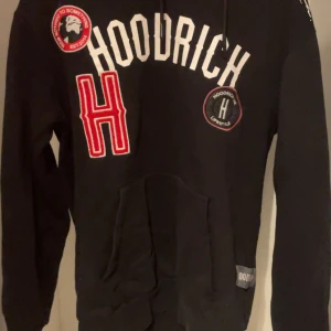 Hoodrich set storlek byxor: M överdel: XS - Svart hoodie från Hoodrich med stora vita och röda tryck framtill. Tröjan har huva med dragsko och en stor magficka. Flera broderade och tryckta detaljer på bröstet och magen. Perfekt för en streetwear-look.