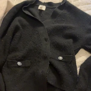 Svart stickad kofta med knappar - Säljer en svart stickad kofta med långa ärmar och knappar framtill. Koftan har två fickor med dekorativa knappar och en klassisk krage. Perfekt att slänga på sig över en t-shirt eller topp.