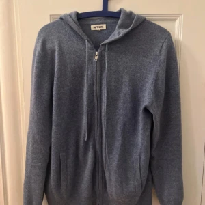 Blå hoodie i kashmir från Soft Goat - Mjuk och lyxig blå hoodie i kashmir från Soft Goat. Tröjan har dragkedja framtill, huva med snören och två fickor. Perfekt för en avslappnad och stilren look.