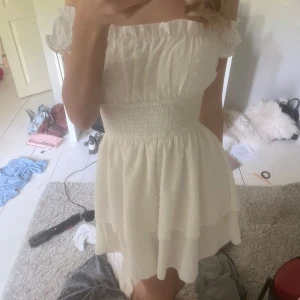 Vit kortklänning med volang och smock - Supersöt vit kortklänning med smock i midjan och volangdetaljer. Klänningen har korta puffärmar och är tillverkad i ett lätt, luftigt material med diskret prickigt mönster. Perfekt för sommarens alla tillfällen!