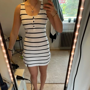 Vit och marinblå randig kortklänning - Säljer en vit kortklänning med marinblå ränder och knappar framtill. Klänningen är ärmlös, har rund halsringning och en liten slits på sidan. Perfekt för varma dagar och enkel att styla med accessoarer.