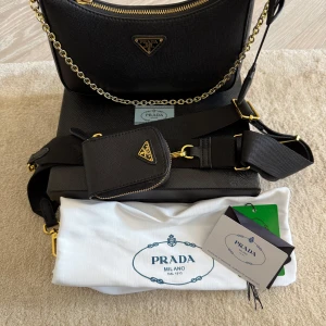 Svart axelväska från Pr*da med gulddetaljer - Säljer en svart axelväska med guldfärgad kedja och detaljer. Väskan har klassisk trekantslogga framtill, justerbar axelrem och medföljande liten plånbok. Kommer med dustbag och kartong. Äkta canvas läder. Köpt för 1900kr