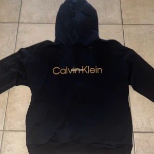 Svart hoodie från Calvin Klein - Säljer en svart hoodie från Calvin Klein med stor gul logga på bröstet. Tröjan har huva och långa ärmar, samt klassisk passform. Perfekt för dig som gillar stilrena märkesplagg. Aldrig använd 