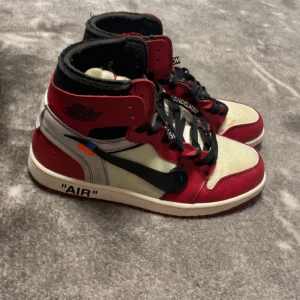 Nike Air Jordan 1 x Off-White Chicago - Säljer ett par Nike Air Jordan 1 x Off-White Chicago med ikoniska röda, vita och svarta paneler, textdetaljer på sidan och extra orange skosnören. Klassisk high-top siluett med svart swoosh och 'AIR'-text på sulan.