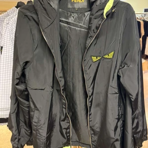 Svart vindjacka från Fendi med huva - Säljer en svart vindjacka från Fendi med huva och dragkedja. Jackan har ett coolt gult och svart ögonmotiv på bröstet och limegrön insida på huvan. Perfekt för dig som vill sticka ut med en exklusiv streetstyle. Alla storlekar finns