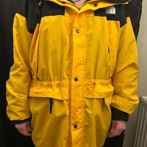 Gul och svart vindjacka från The North Face - Säljer en gul och svart vindjacka från The North Face med huva och dragkedja samt knappar framtill. Jackan är i två delad och har justerbar midja och flera fickor med dragkedja. Uppskattad storlek är som XL. Perfekt för blåsiga dagar och har en sportig look.