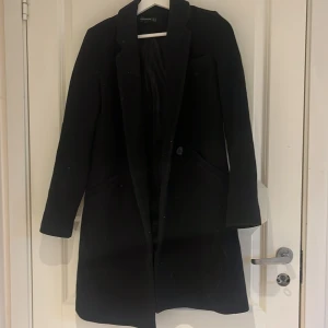 Svart kappa - Stilren svart kappa med klassisk krage och knäppning framtill. Lång modell med diskreta fickor på sidorna. Perfekt för en snygg och tidlös look.
