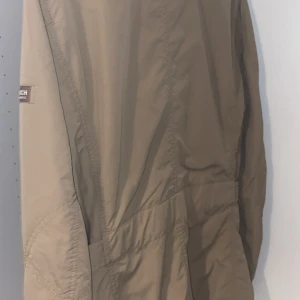  Woolrich safarijacka  - Säljer en beige jacka från Woolrich med fyra stora fickor framtill och knappar i silverfärg. Jackan har hög krage med spänne och lång ärm. Perfekt för dig som vill ha en stilren och funktionell jacka.